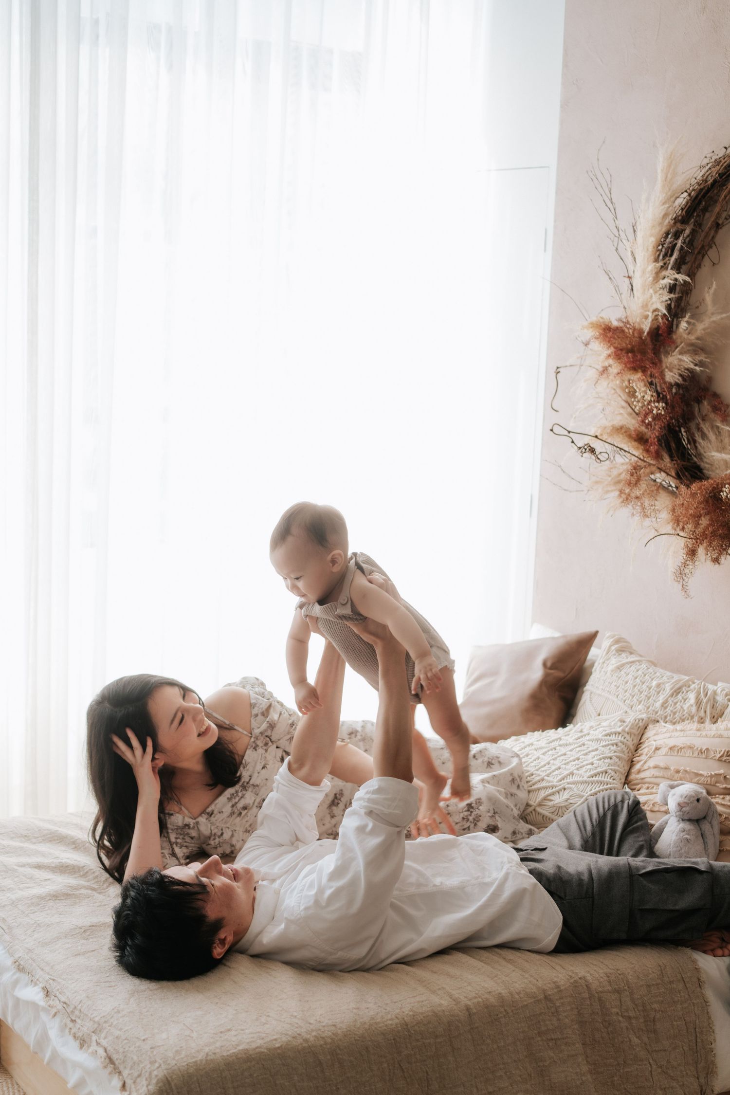 A parent and baby share a tender moment while lying together on a brown couch in a bright room.