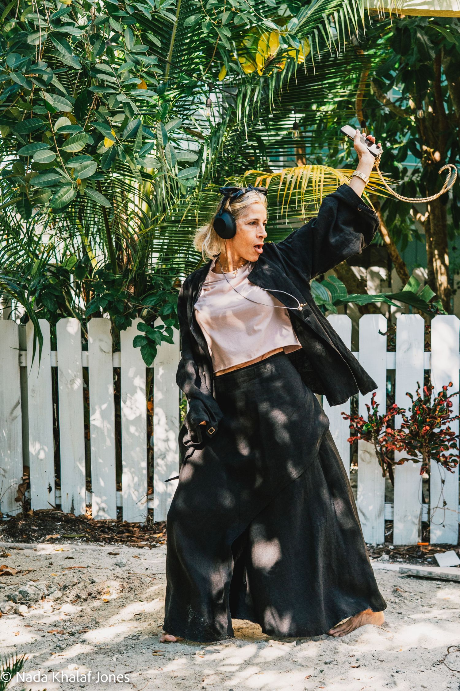 A person in a dress sits next to a large black dog on a sandy ground with tropical foliage and white fence in background.