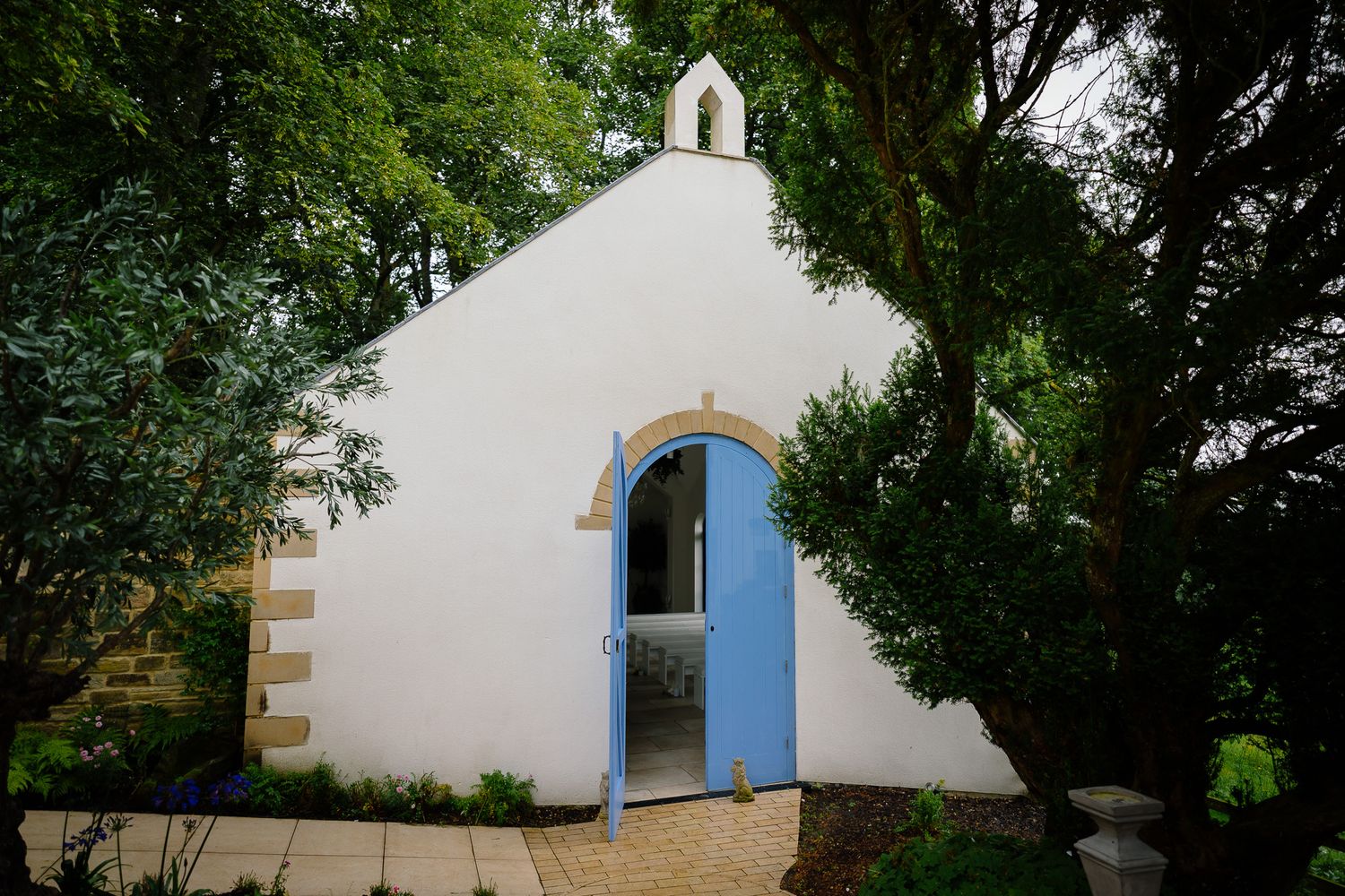 White chapel exterior with blue door surrounded by green trees and landscaping. Woodhill Hall.