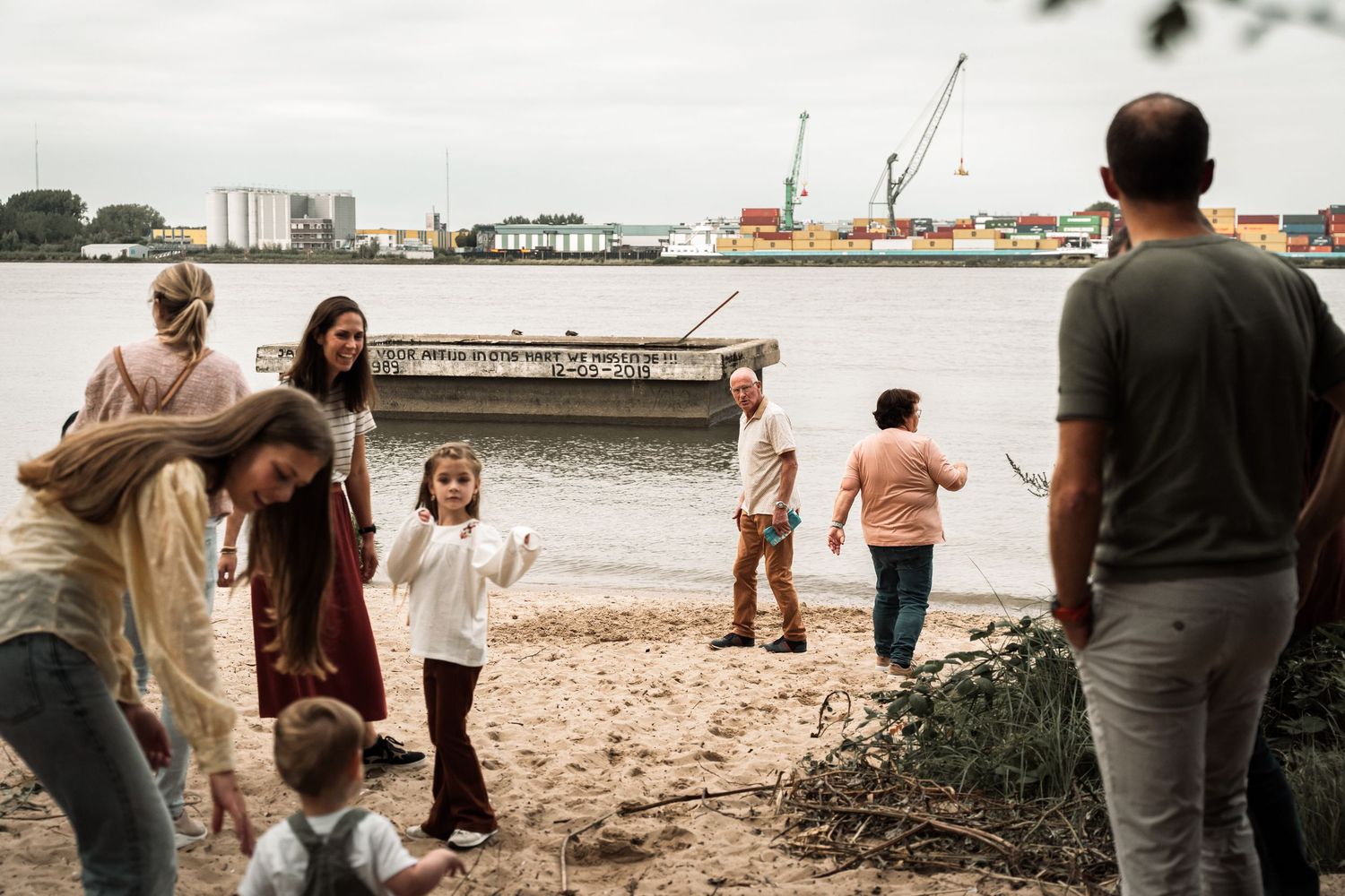 People gather along a beach shoreline with boats and buildings visible in the background on an overcast day.