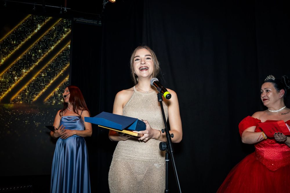 Young woman in silver dress speaking at microphone on stage at awards ceremony.