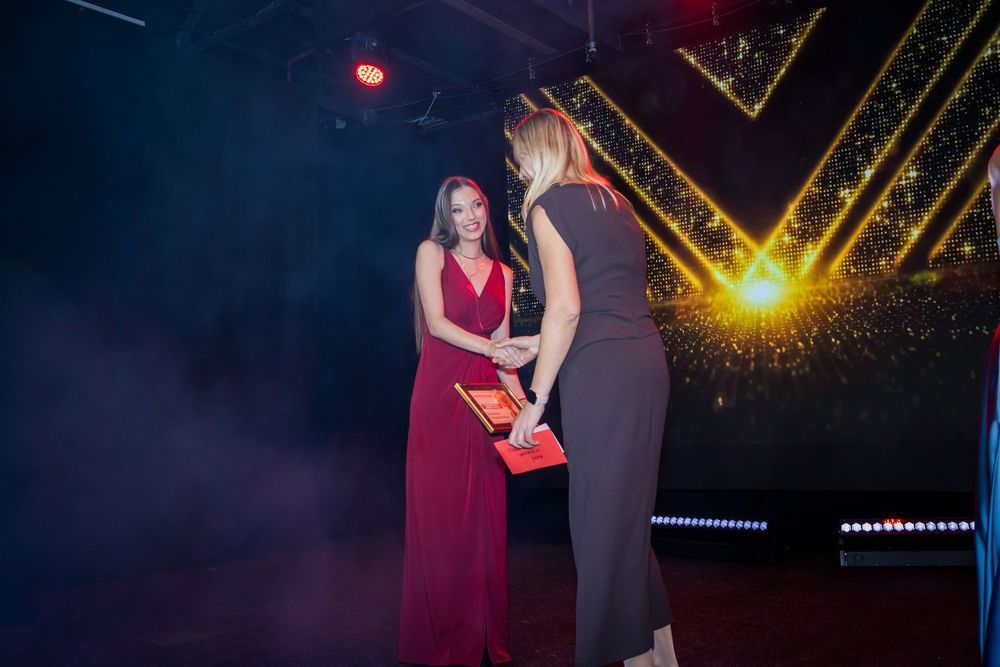 Two women in formal gowns exchange award plaque on dramatic fog-filled stage.