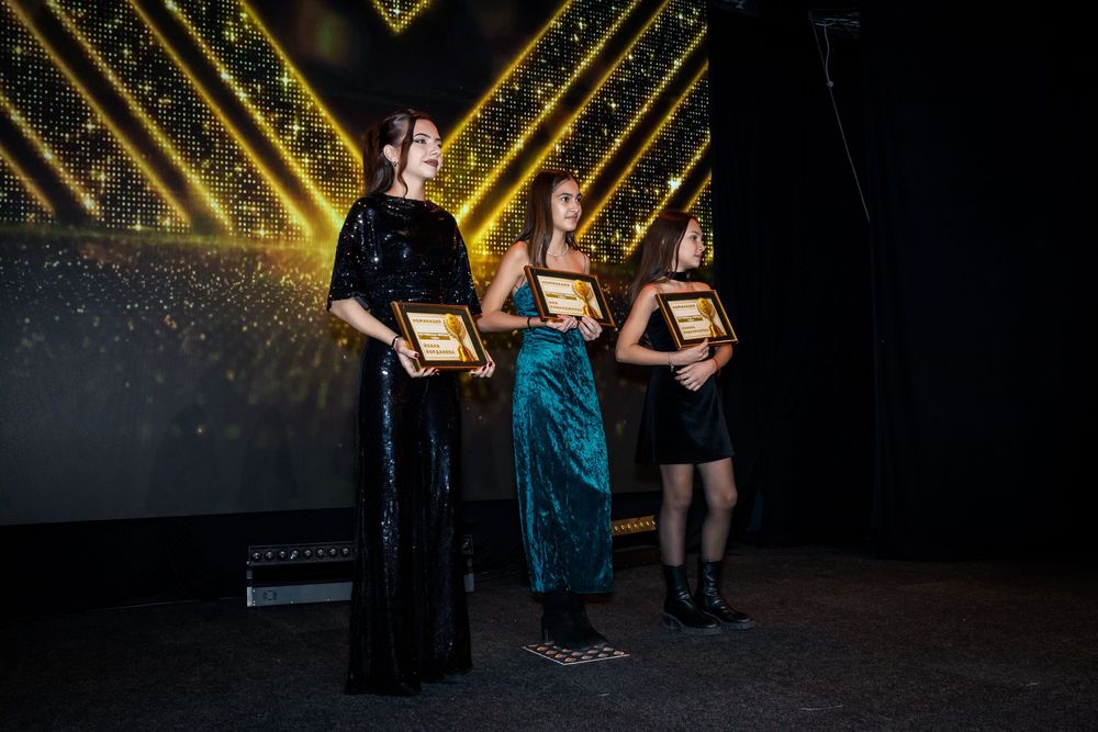 Three elegantly dressed women holding golden plaques on stage at awards ceremony.