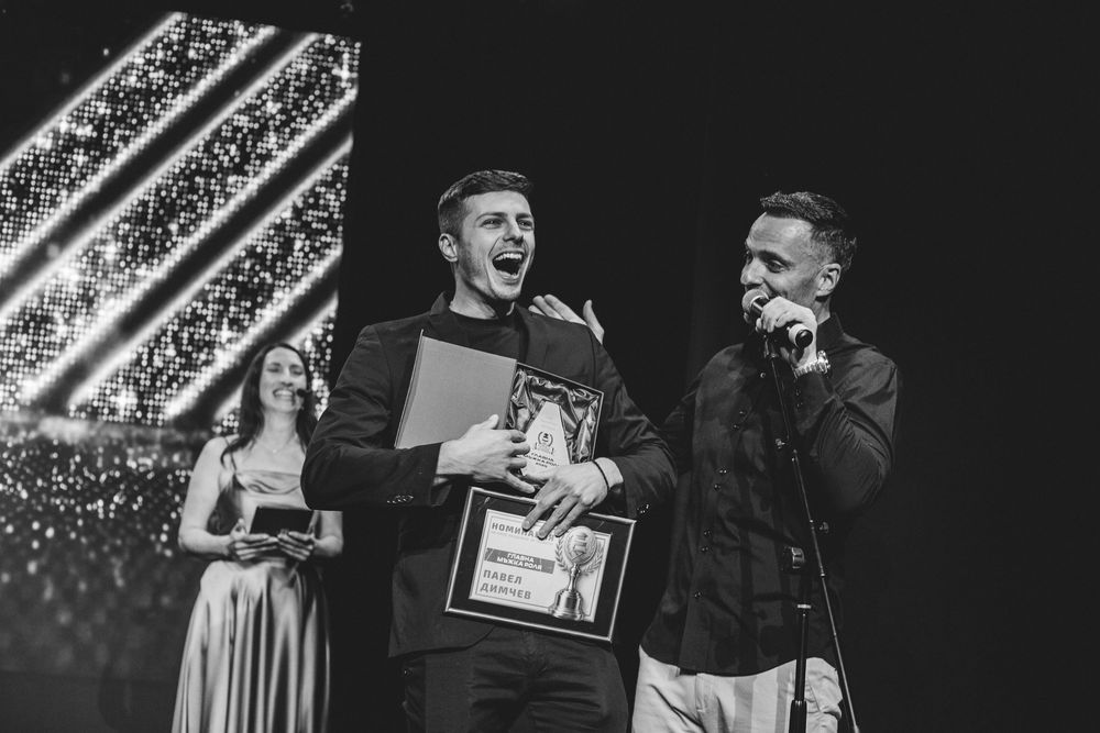 Man smiling on stage holding award at black and white ceremony event.