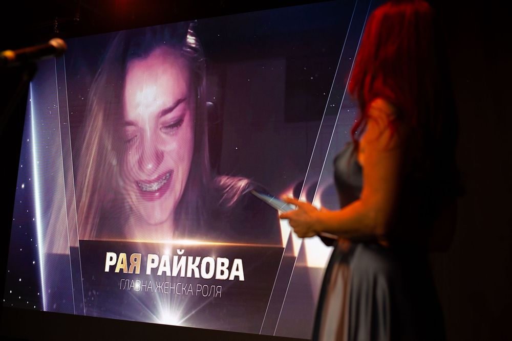Presenter standing beside large screen displaying nominee Raya Raykova at awards show.