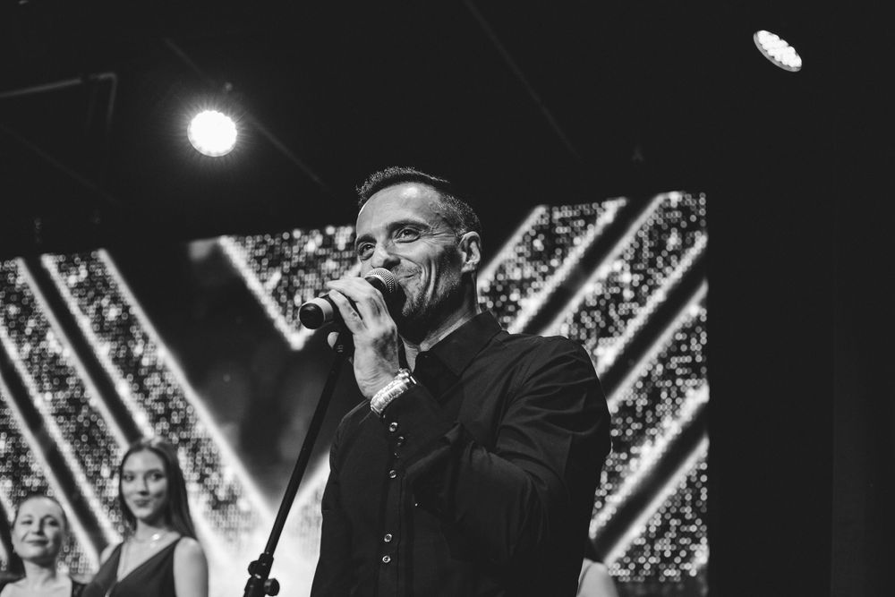 Man in black suit speaks into microphone on stage at an elegant awards ceremony with bokeh lights.