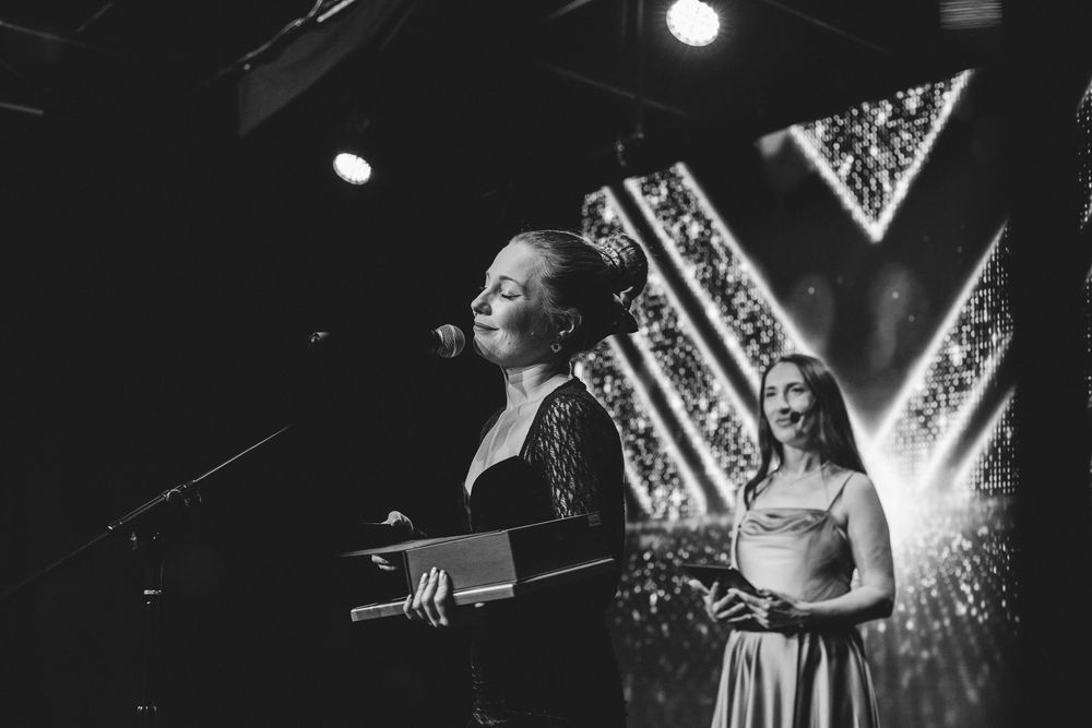 Two women stand at podium on stage during black and white awards ceremony with sparkling background.