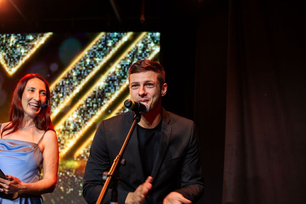 Young man in black blazer speaks at microphone beside woman in blue dress at awards ceremony.