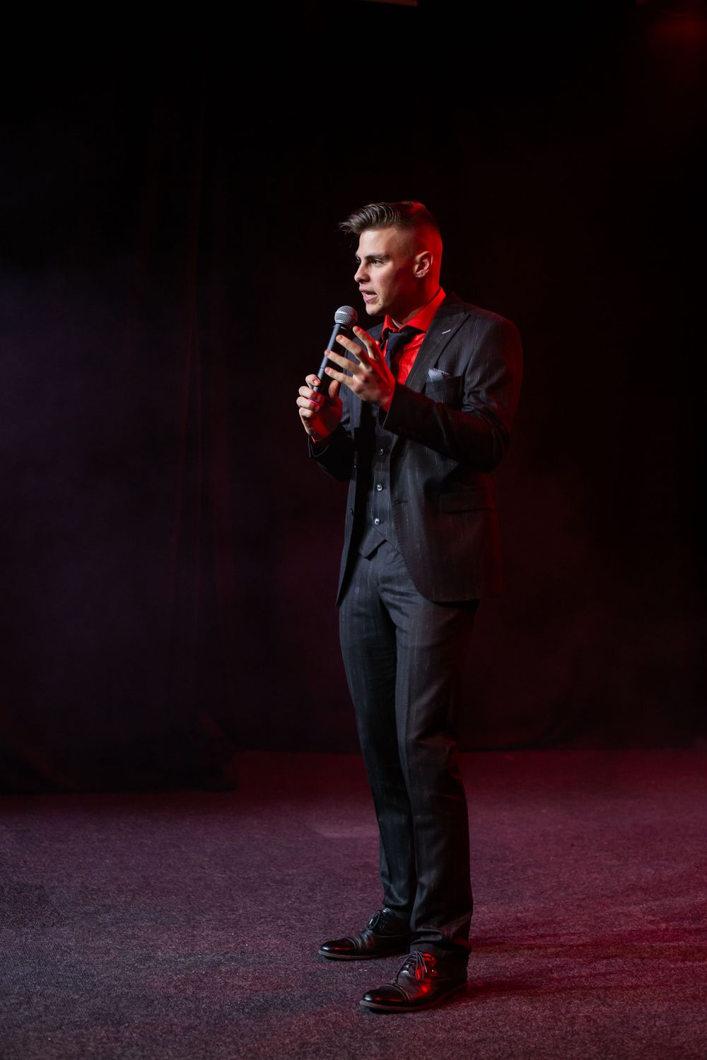 Young man in black suit holds microphone on stage under dramatic red lighting.