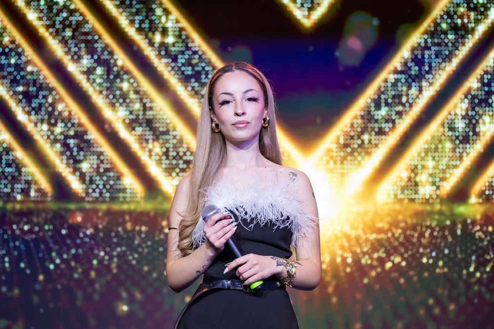 Elegant blonde woman in black and white feathered dress stands before glittering stage backdrop.