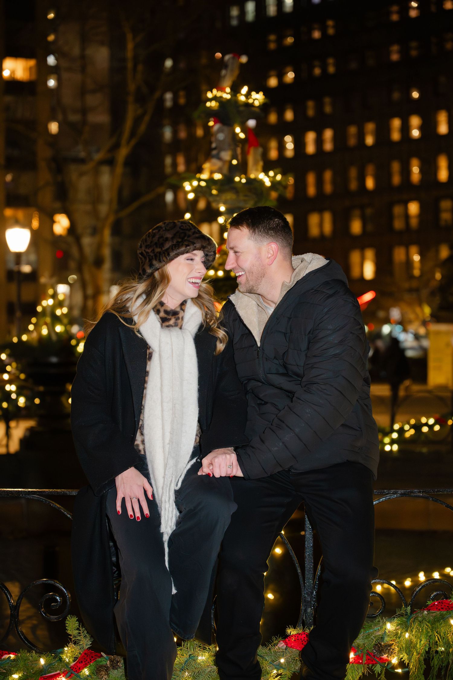 A newly engaged couple shares a romantic moment on a park bench surrounded by holiday lights and decorations at night in Madison Square Park