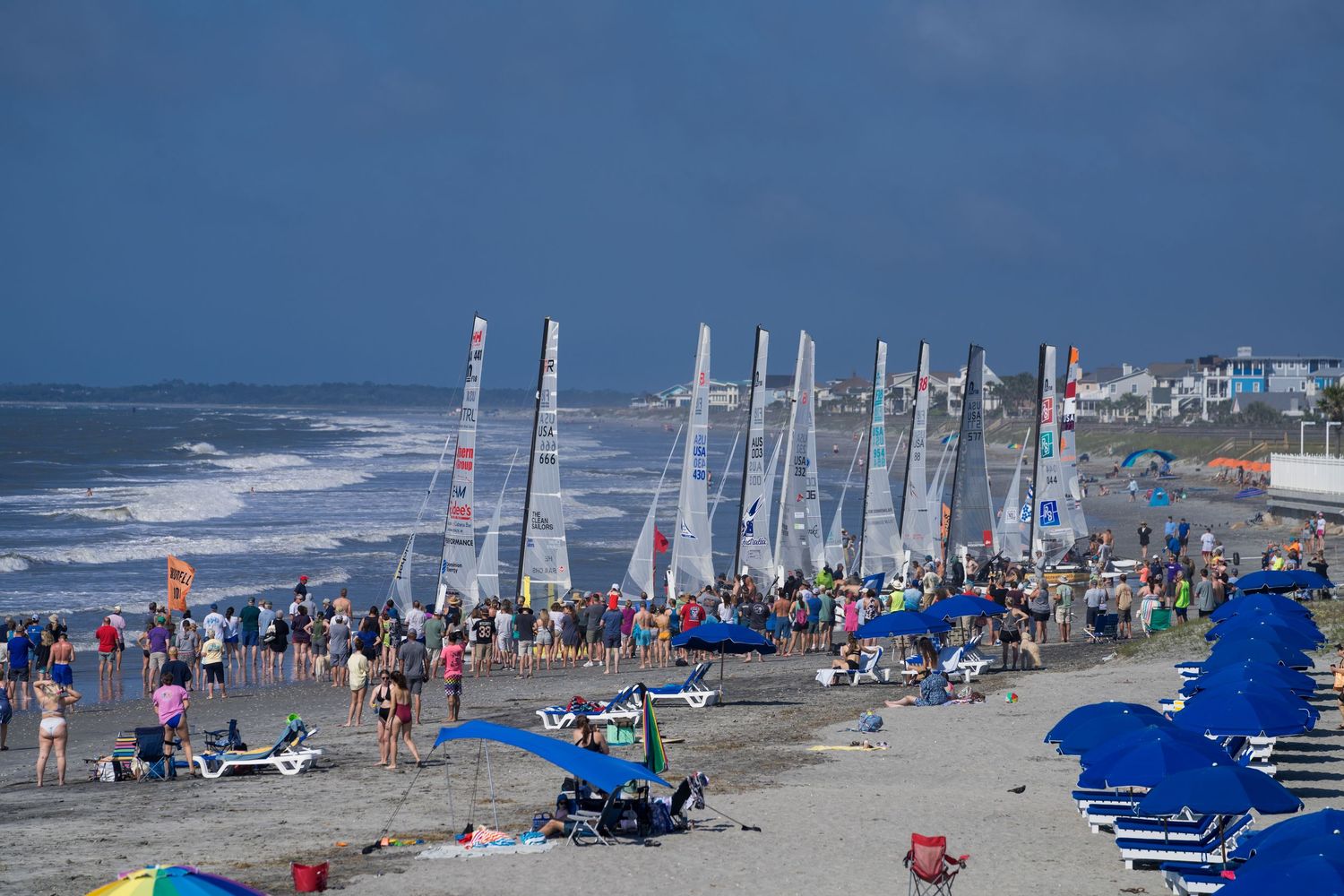 Colorful sailboats and beach chairs line a sunny oceanfront beach with dark storm clouds approaching in the distance.