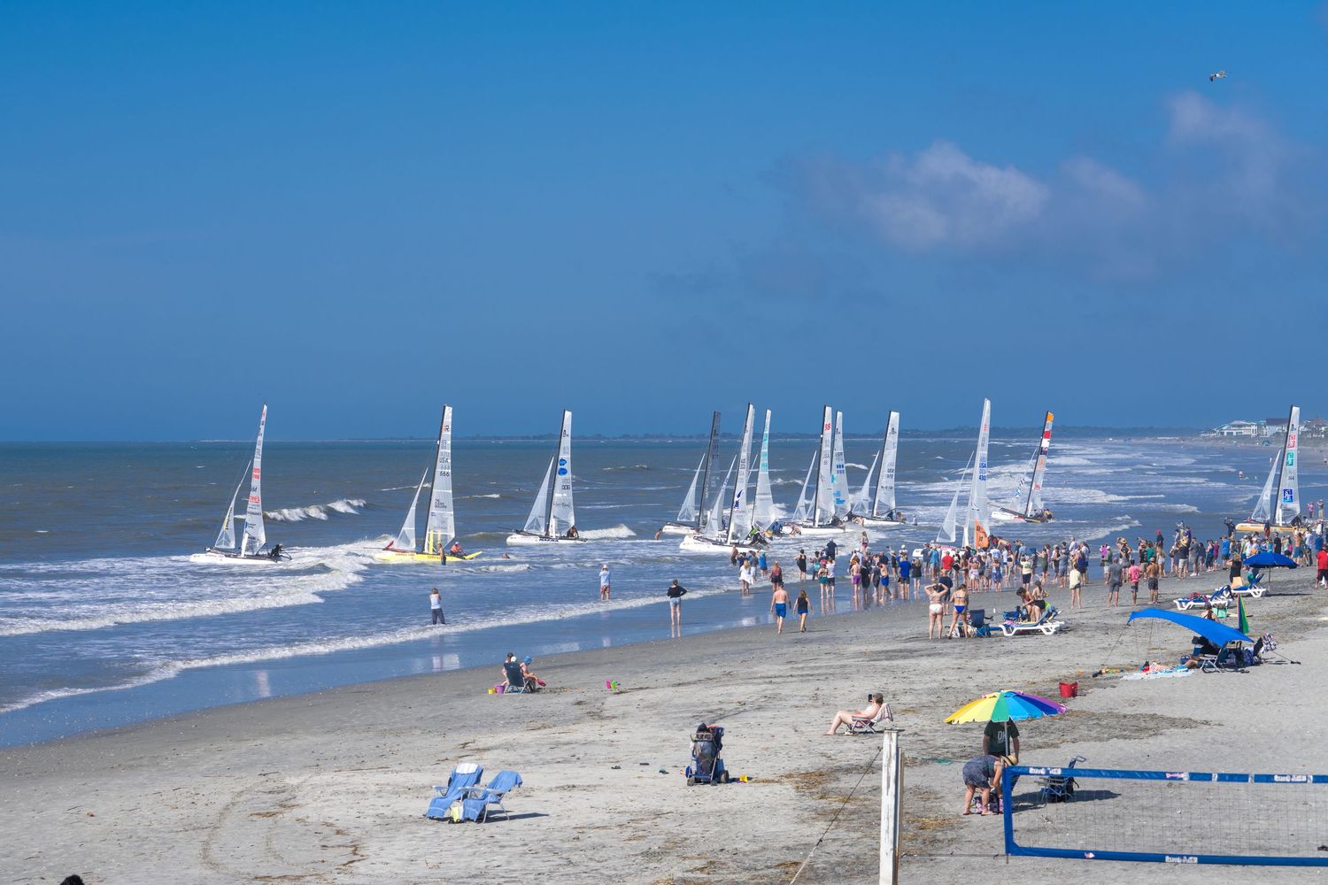 Sailboats line up along a sunny beach with blue umbrellas and beachgoers enjoying the sandy shoreline.