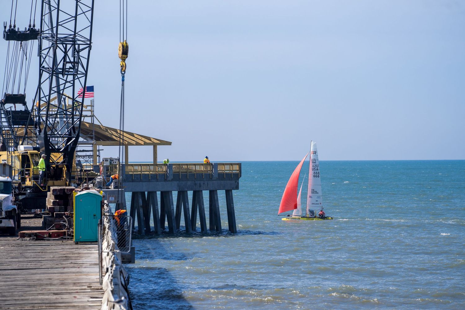 Construction cranes and equipment line an industrial pier extending into the blue ocean waters on a sunny day.