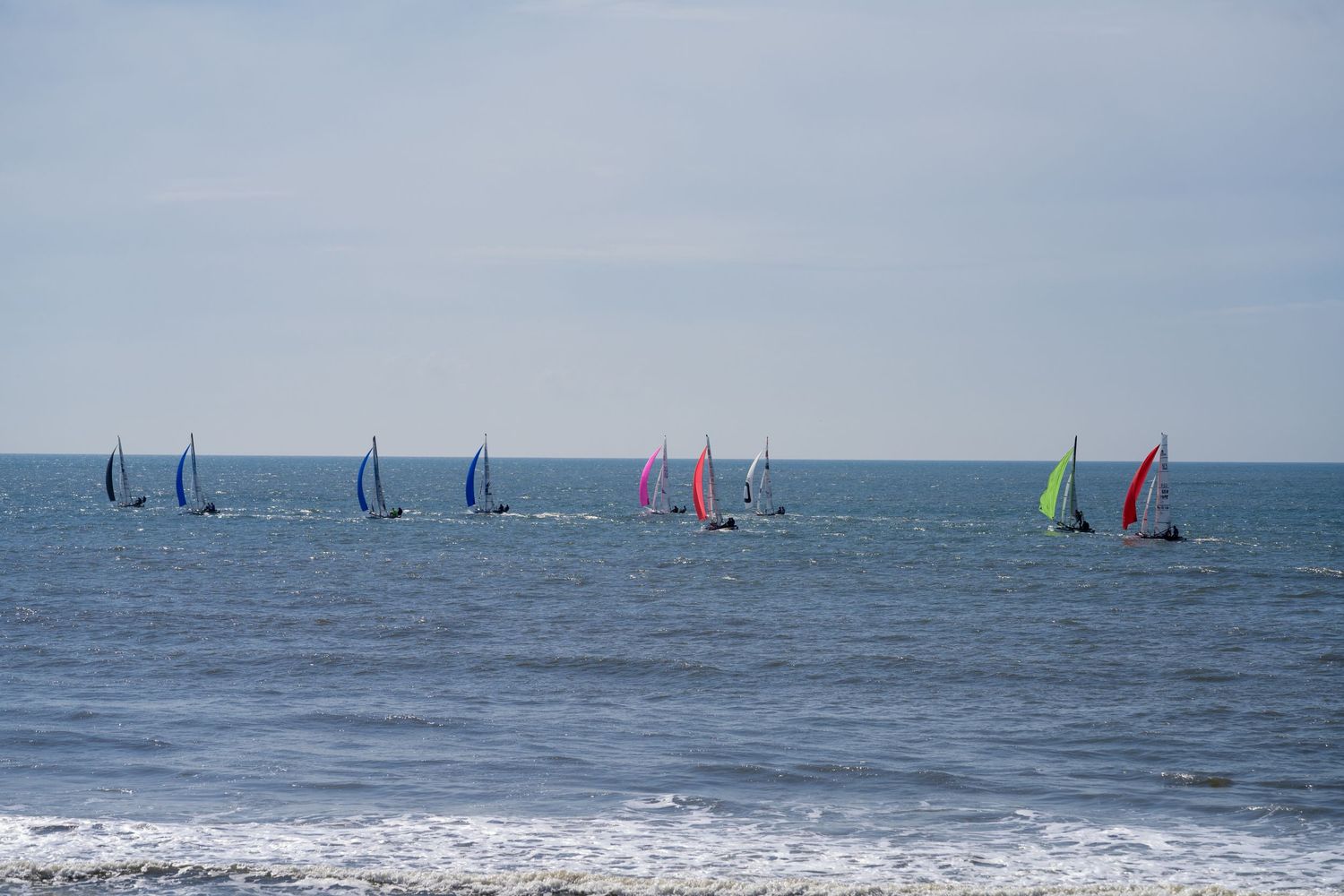Colorful sailboats with vibrant spinnaker sails race across blue ocean waters on a cloudy day.