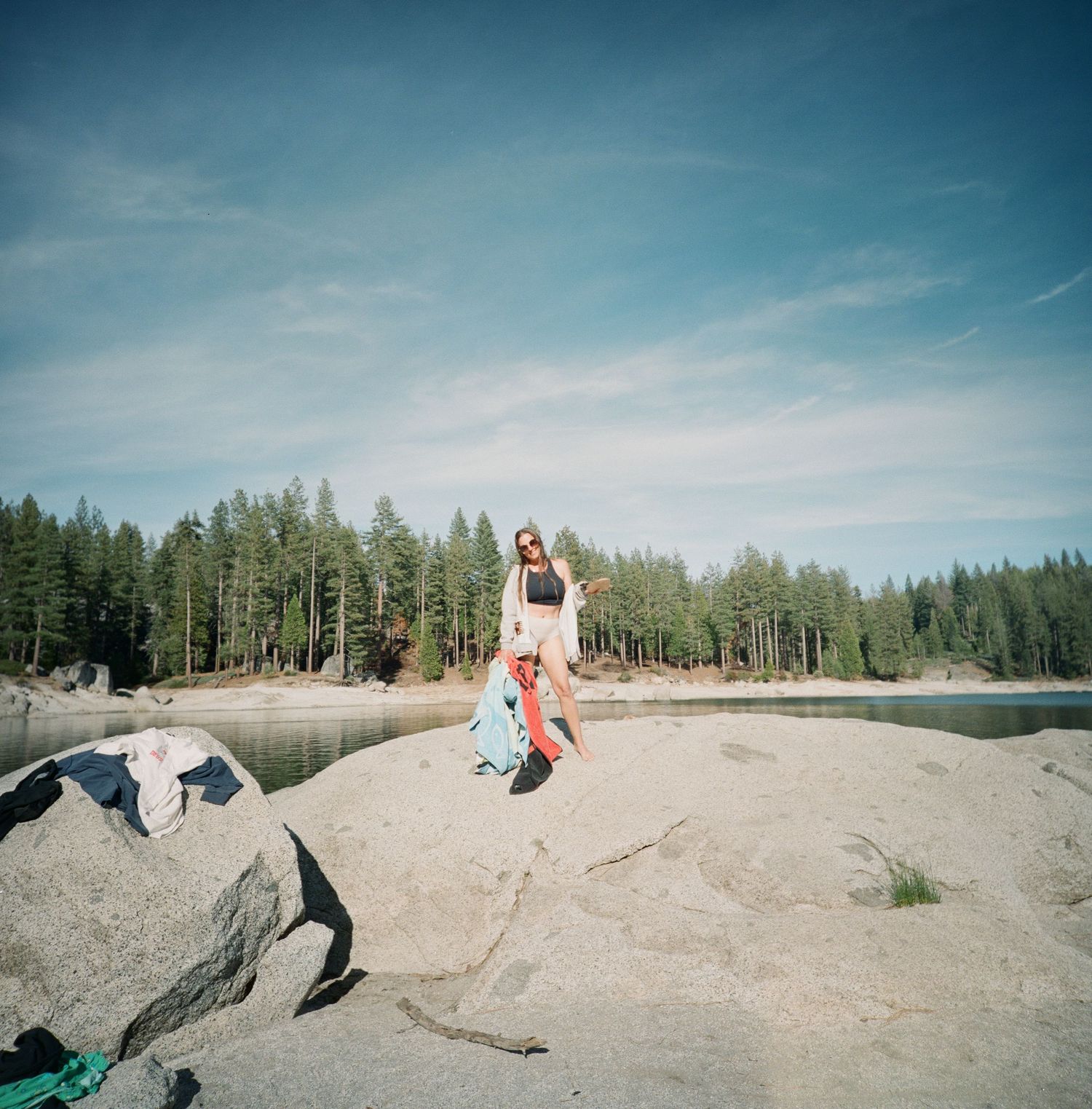 A sequence of people sliding down a sandy slope in a forested area near a lake on a sunny summer day.
