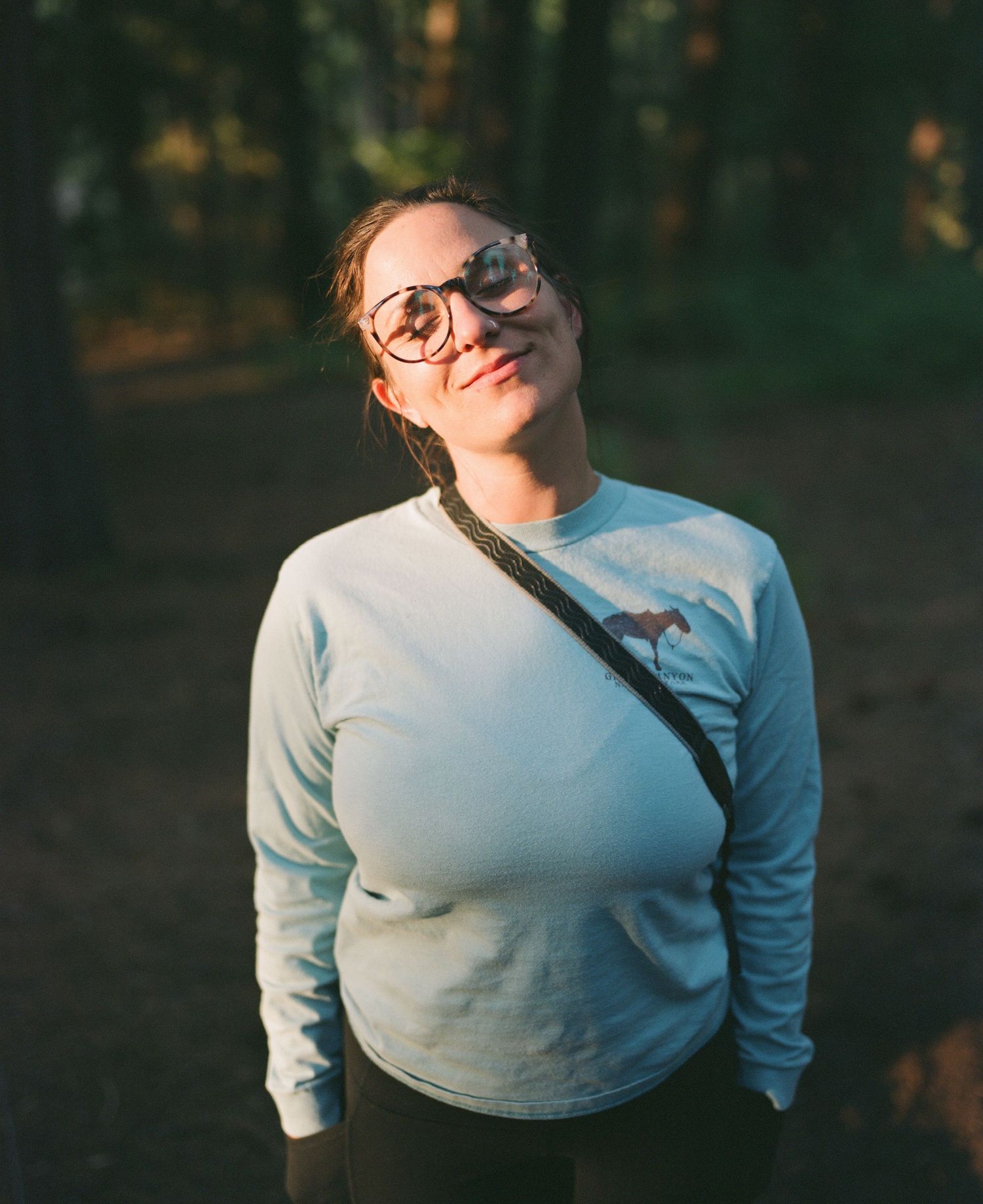 Person in white sweater poses playfully against dark forest background during sunset.