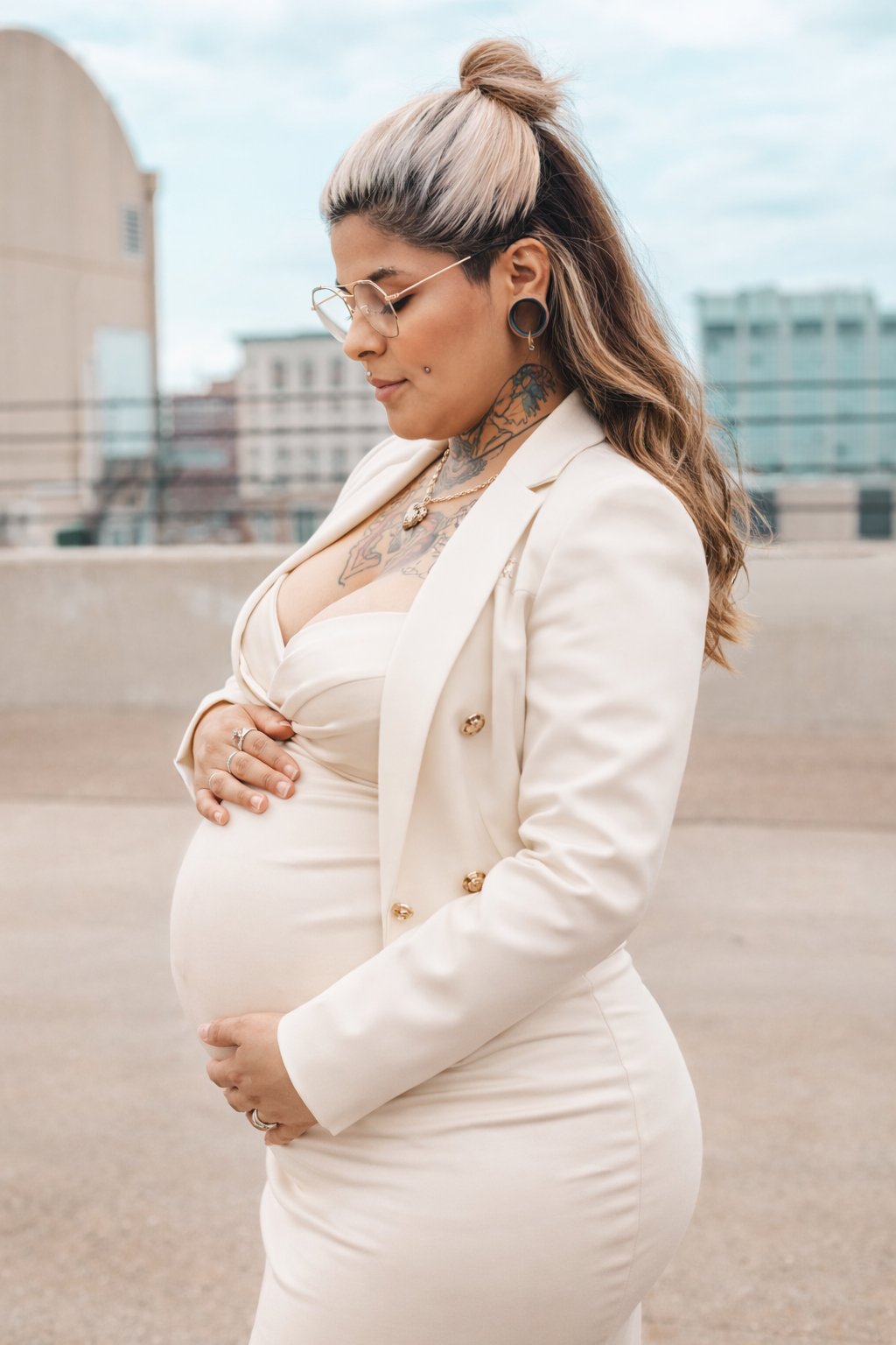 A pregnant woman in a white blazer poses outdoors against an urban backdrop in a maternity photoshoot series.