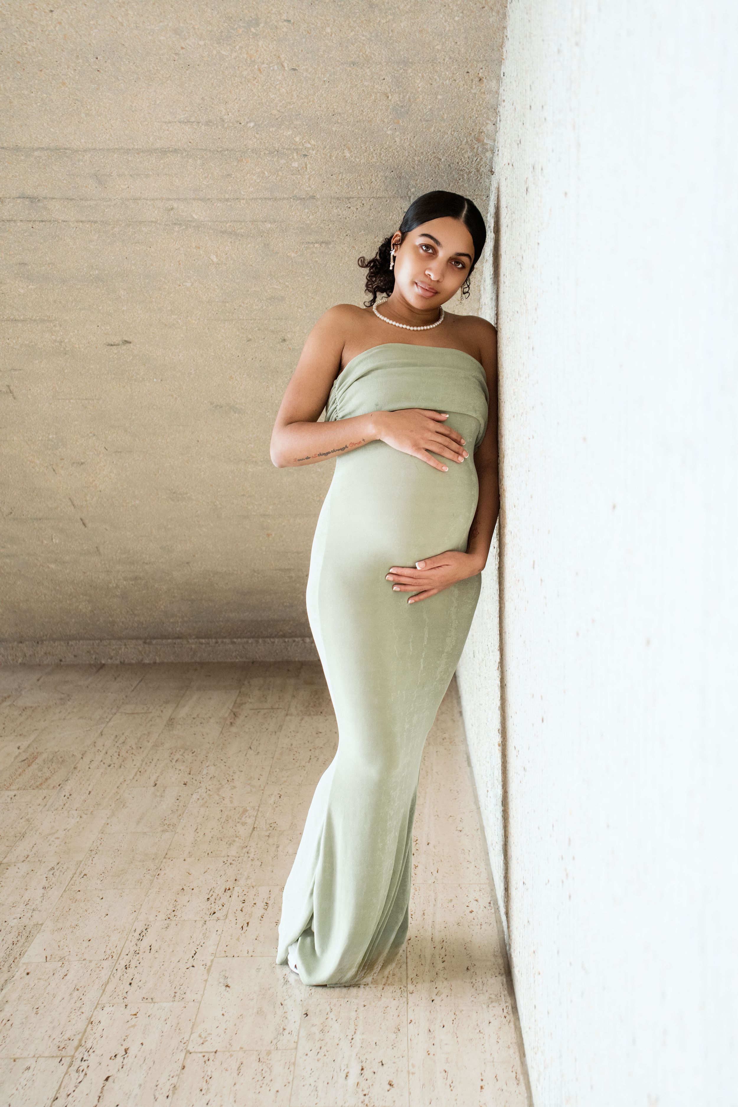 A person in a mint green dress poses elegantly against a light-colored wall in a minimalist setting.
