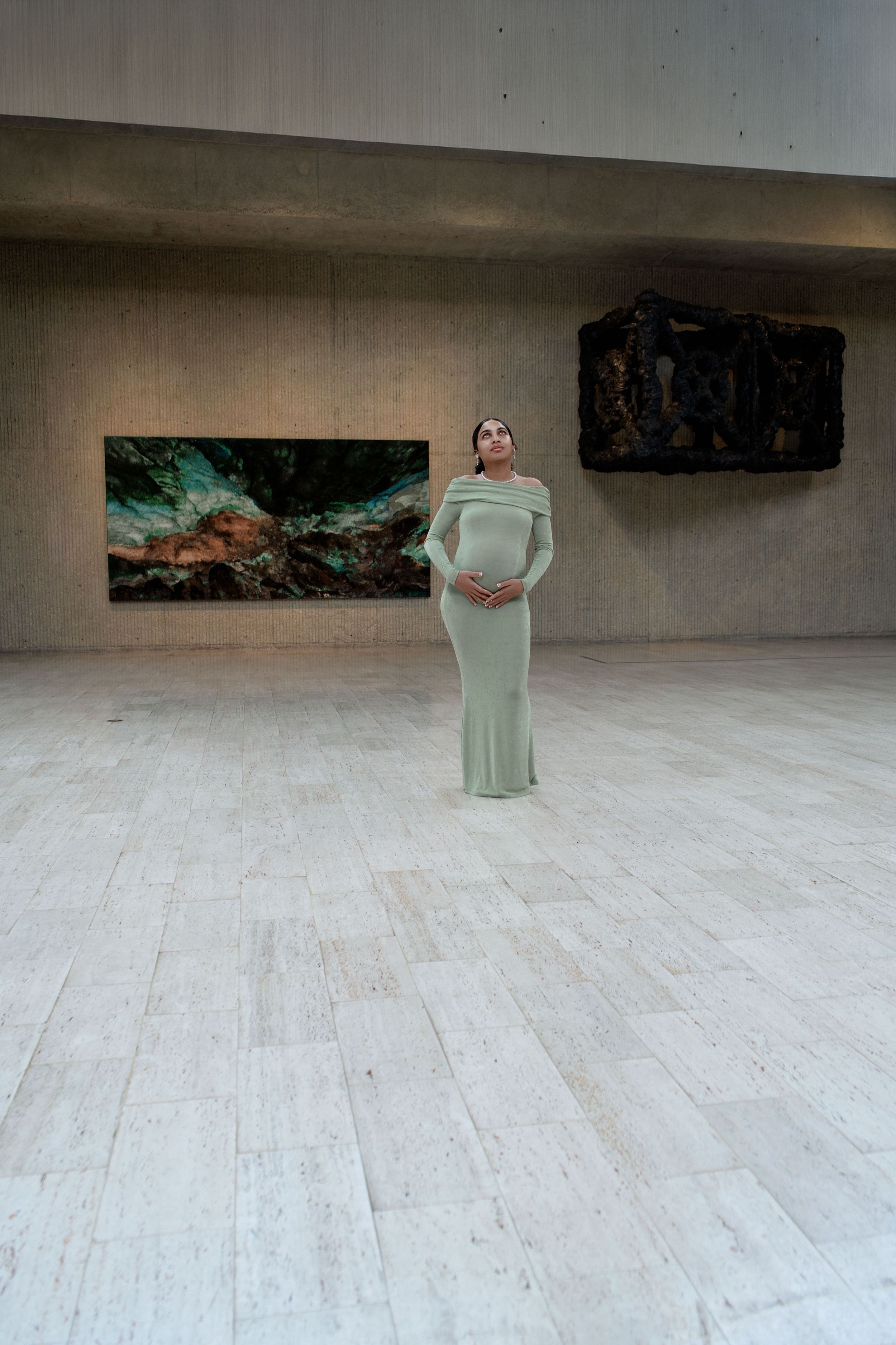 Figure in a sage green dress performs a contemporary dance sequence in a minimalist gallery space with wooden floors.