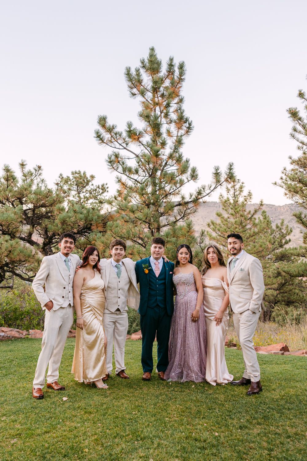 Wedding party in elegant neutral tones poses outdoors with pine trees and mountains in the background.