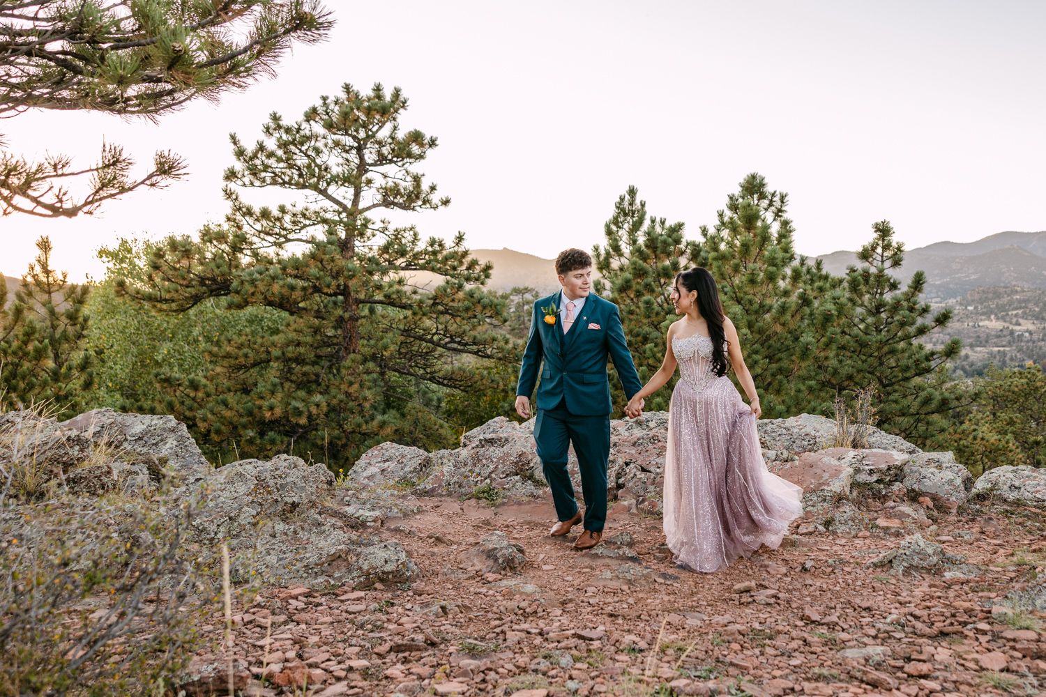 Couple in formal attire sits on rocky mountain ledge overlooking scenic mountain landscape at sunset.