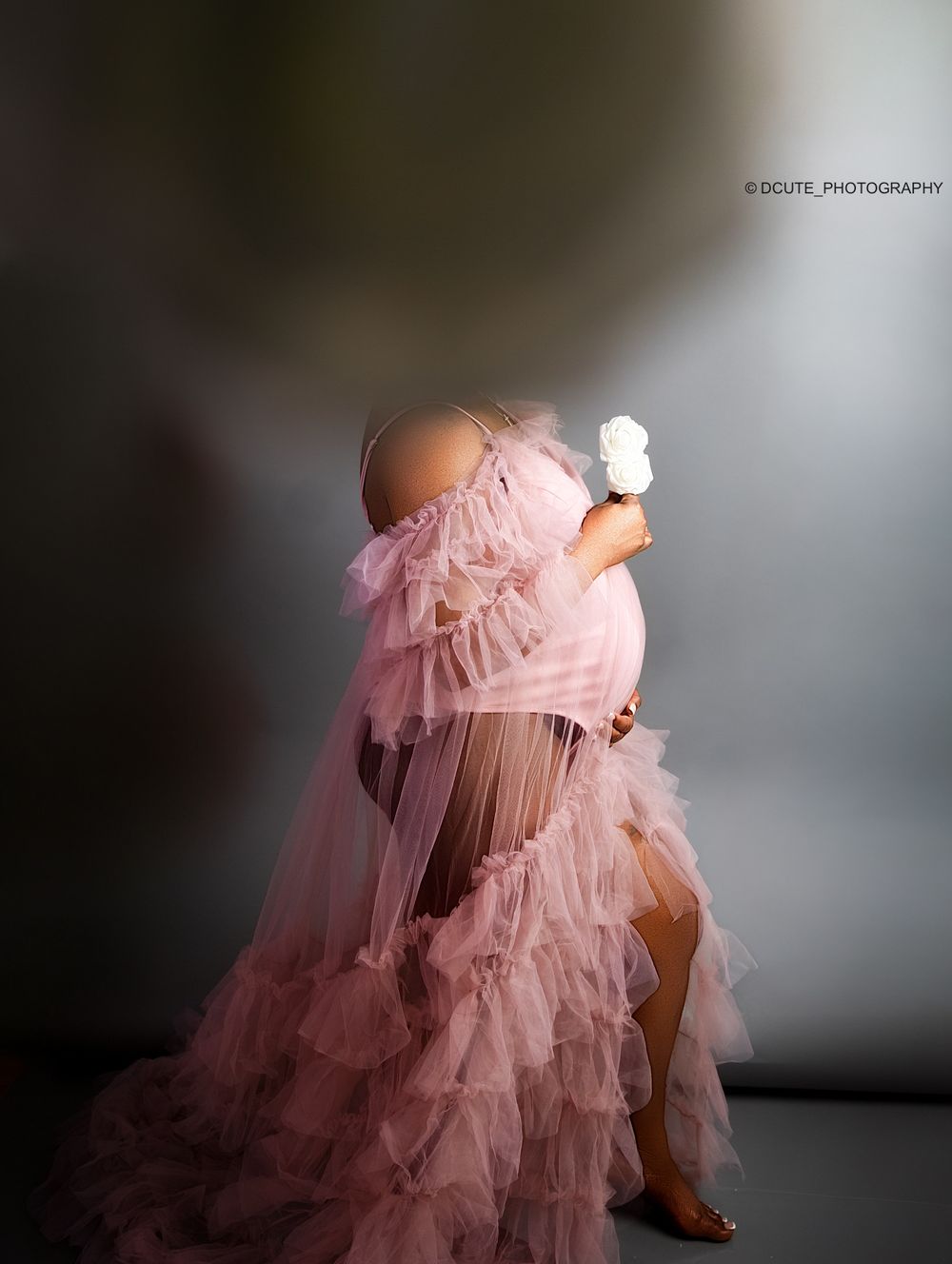 Pregnant woman in a flowing pink tulle maternity gown holding a white rose in a professional studio photo.