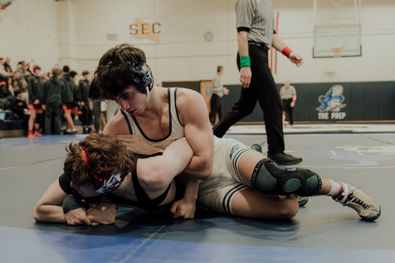 High school wrestlers compete in a wrestling match on a gymnasium mat with spectators watching in the background.