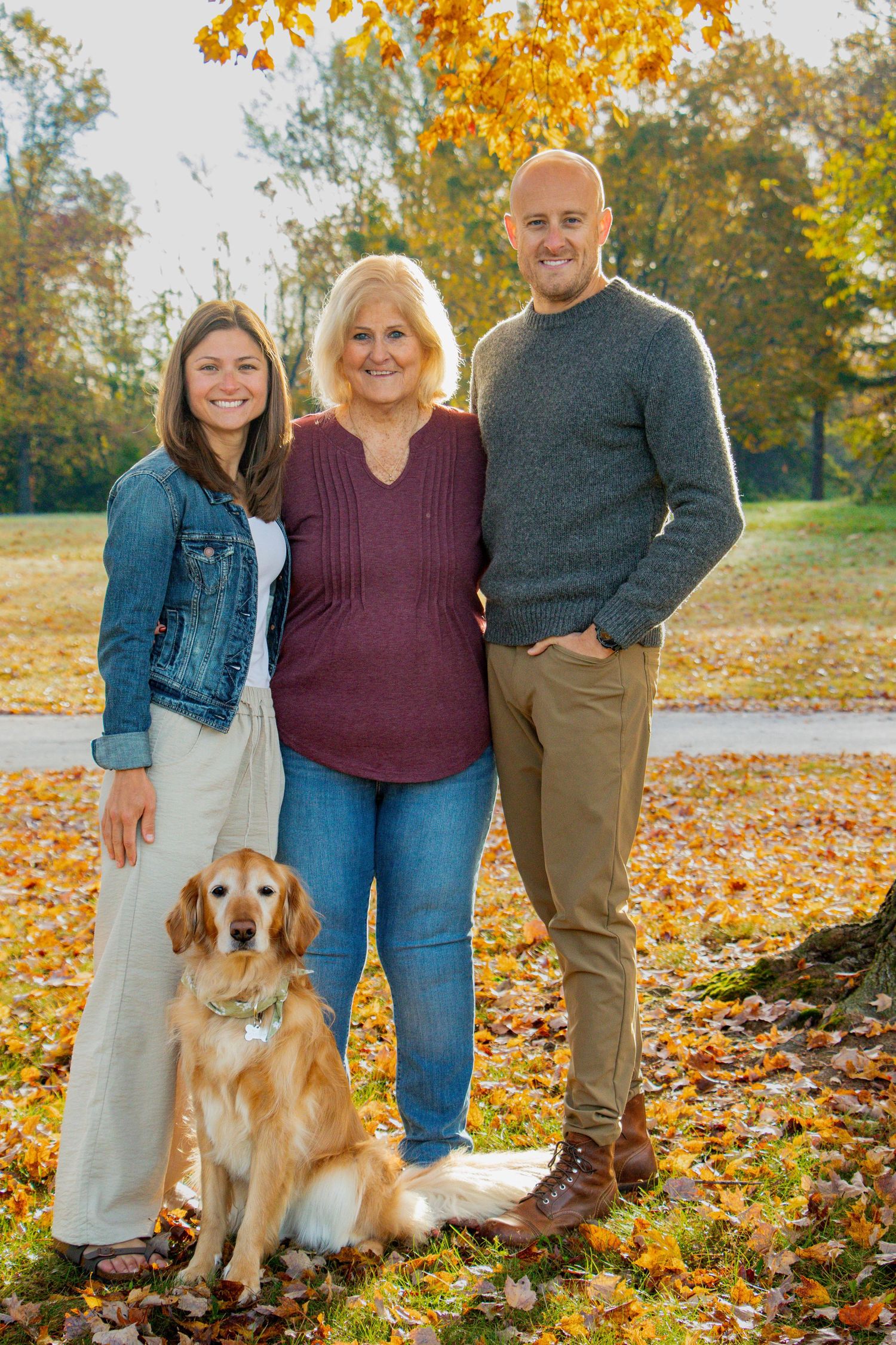 Two adults and a Golden Retriever posing together in a park with colorful fall foliage.