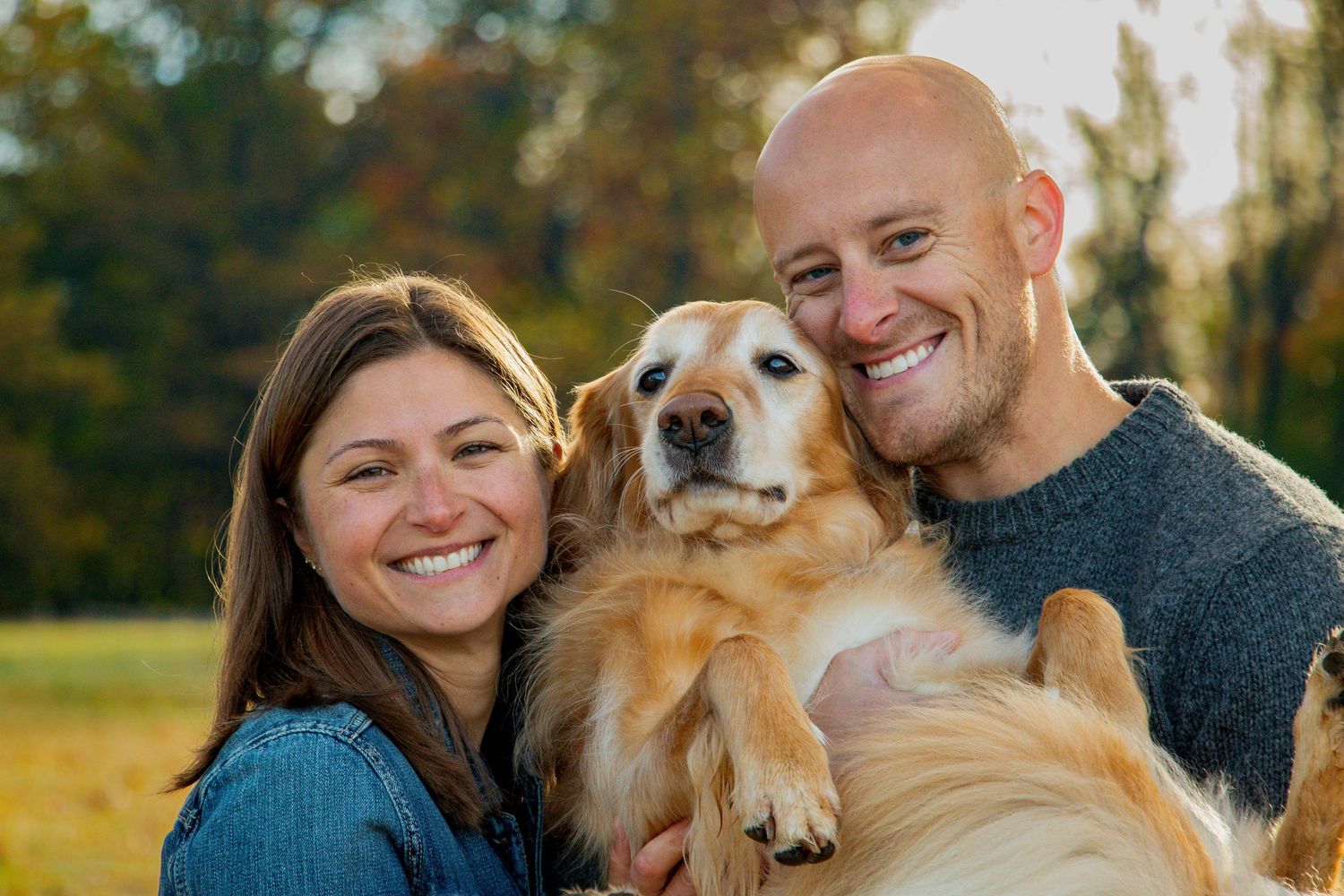 Smiling couple holds their Golden Retriever dog outdoors during a warm autumn day photo session.
