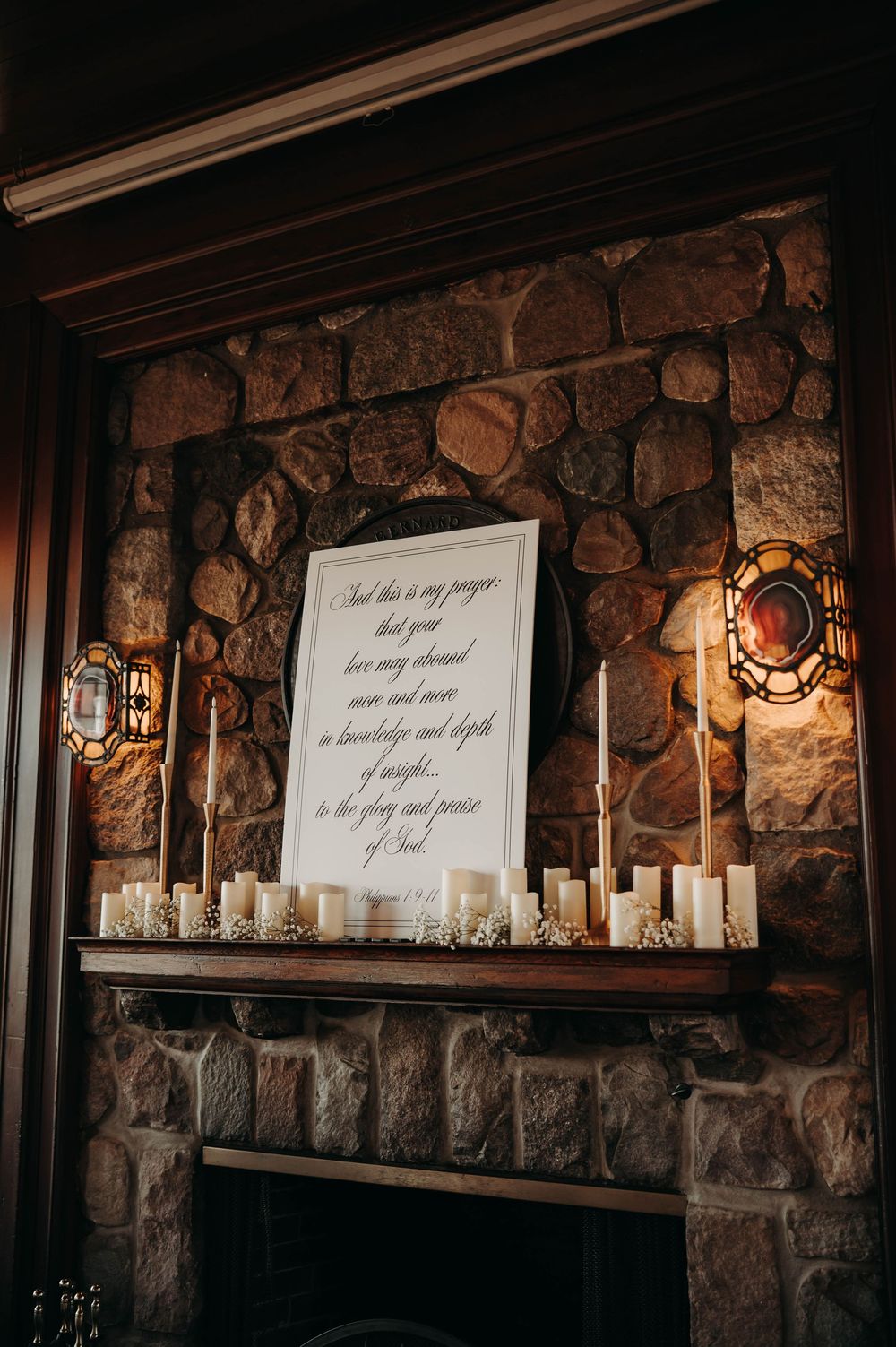 Rustic stone fireplace mantel adorned with white candles and a framed calligraphy prayer sign at a wedding venue.