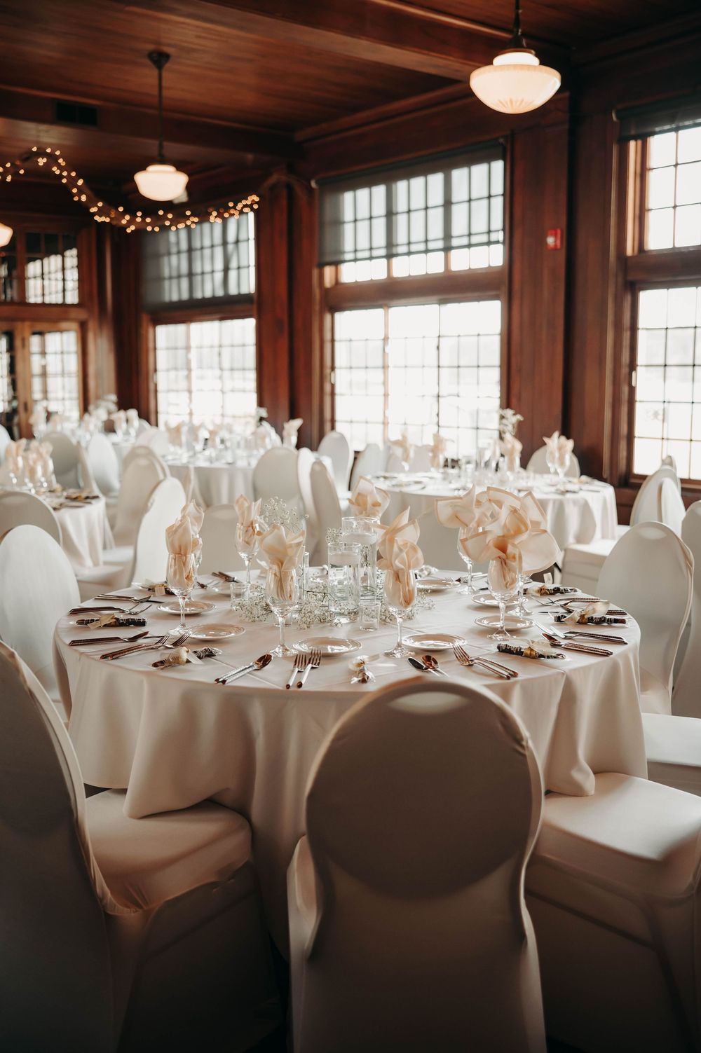 Sunlit wedding reception hall with white round tables, ghost chairs, and large windows in background.