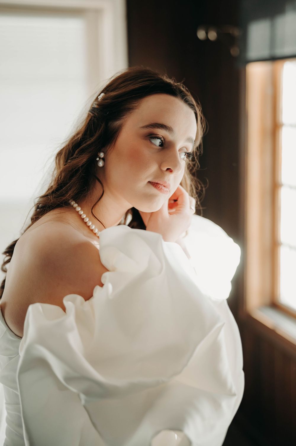 Beautiful bride in white off-shoulder gown wearing pearl jewelry gazing thoughtfully near a sunlit window.