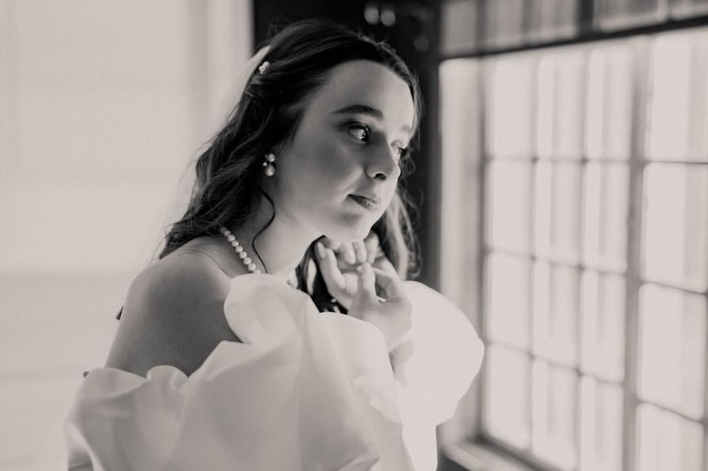Black and white portrait of a bride in an off-shoulder gown wearing pearls near a window.