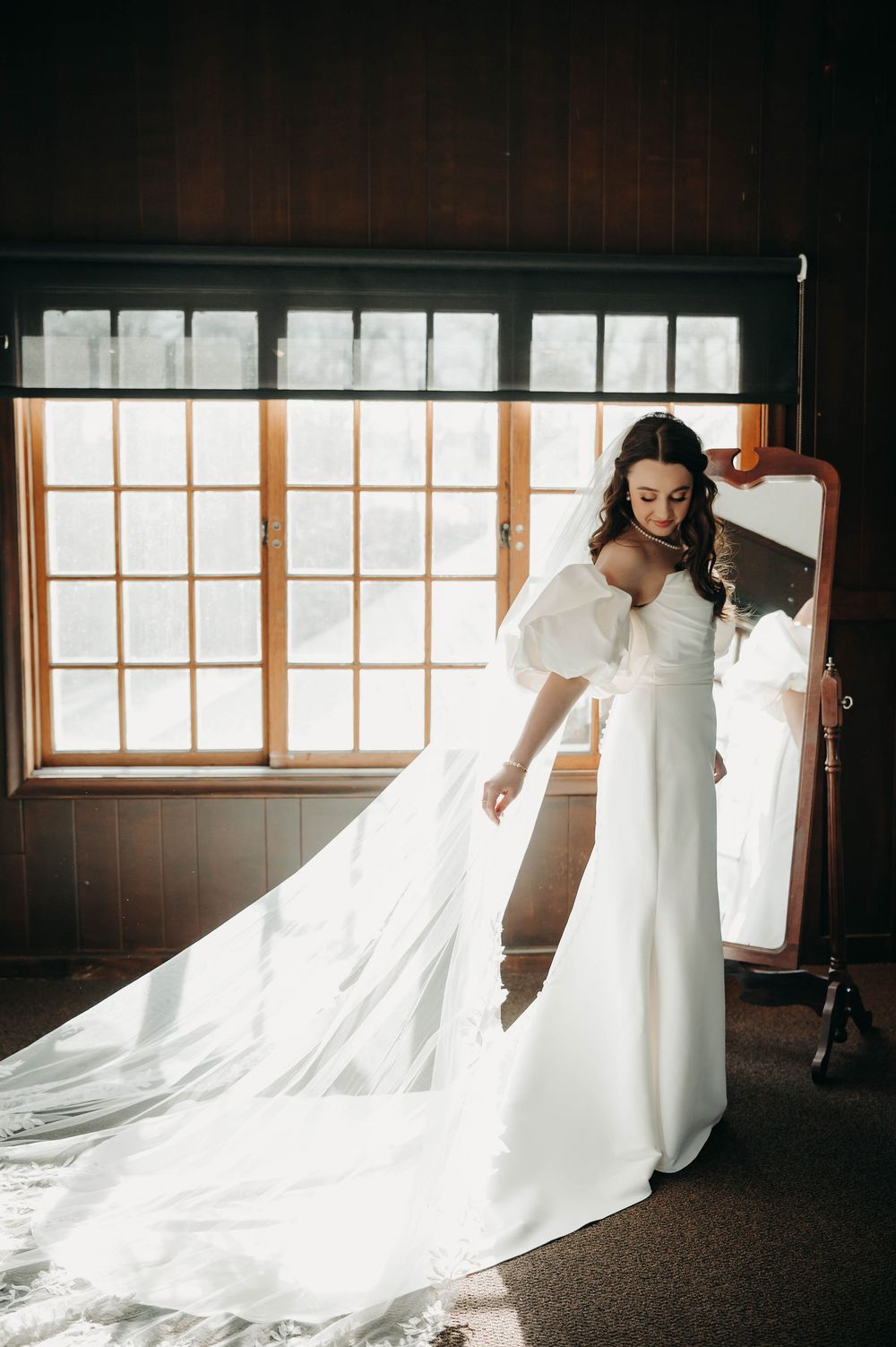 Bride in white off-shoulder gown with puff sleeves adjusts her cathedral veil beside a vintage mirror.