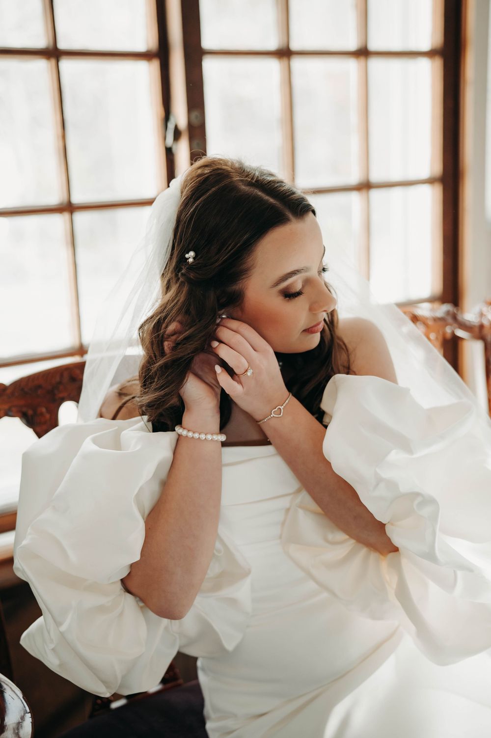 Bride in white puff-sleeve gown adjusts her earring while seated near a sunlit window on her wedding day.