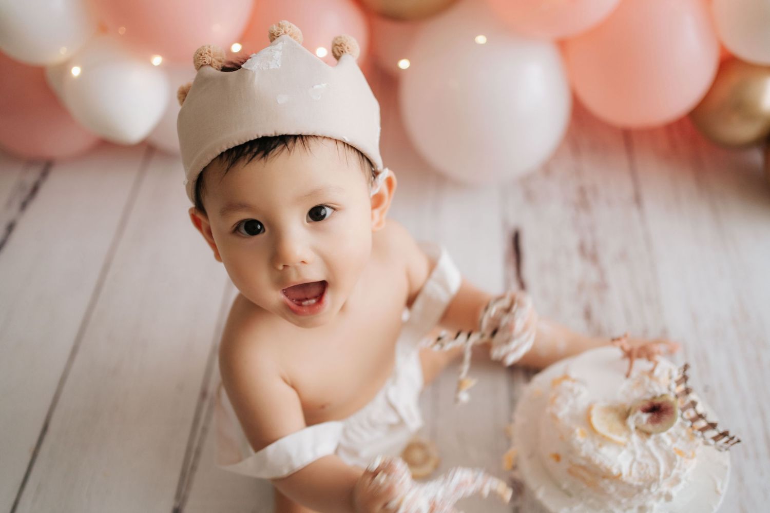 A baby sits in a white chair surrounded by floral decorations and balloons at a birthday celebration.