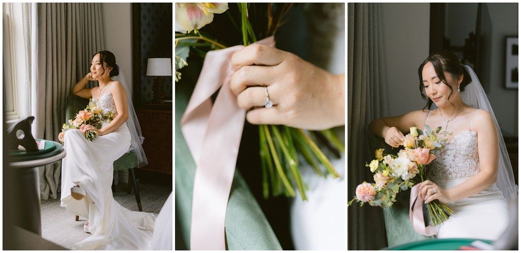 A bride in a white lace gown holds a bouquet while getting ready in a softly lit room.