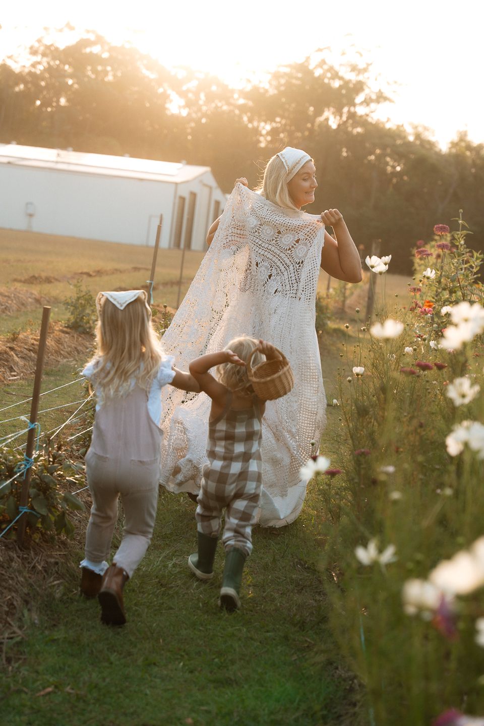 Two people in white dresses walk through a dreamy wildflower field at golden hour as sunlight filters through the scene.