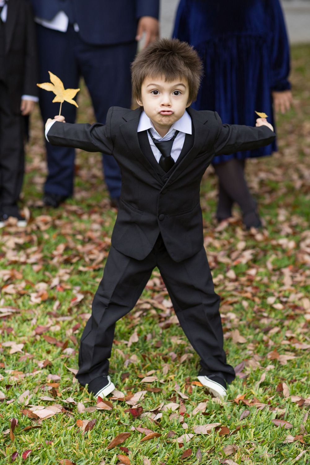 Young ring bearer in black suit poses playfully with fallen leaves.
