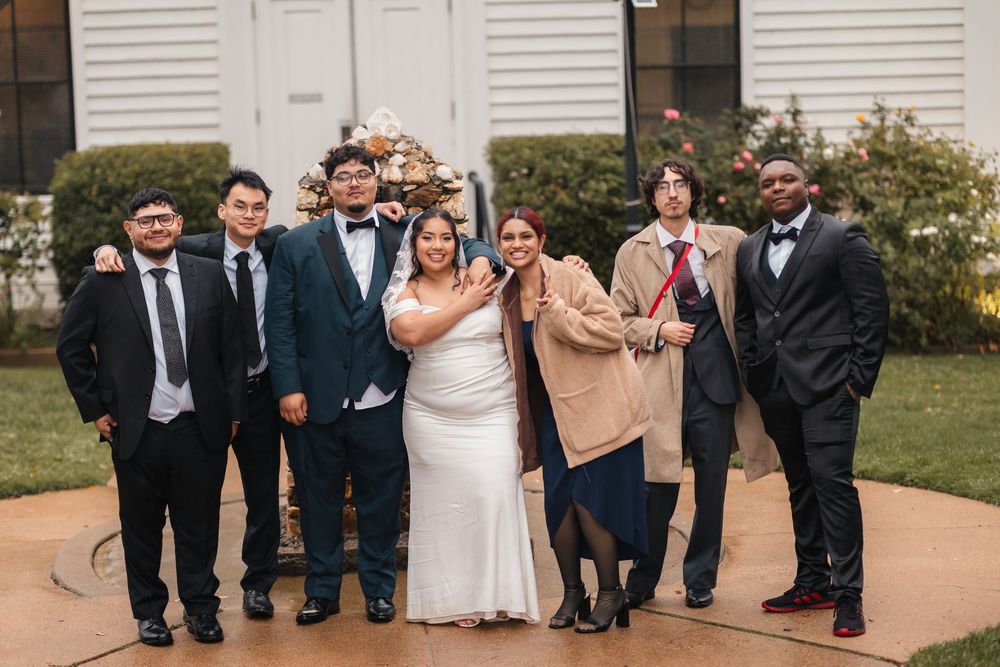 Wedding party poses together on concrete path in front of white building.