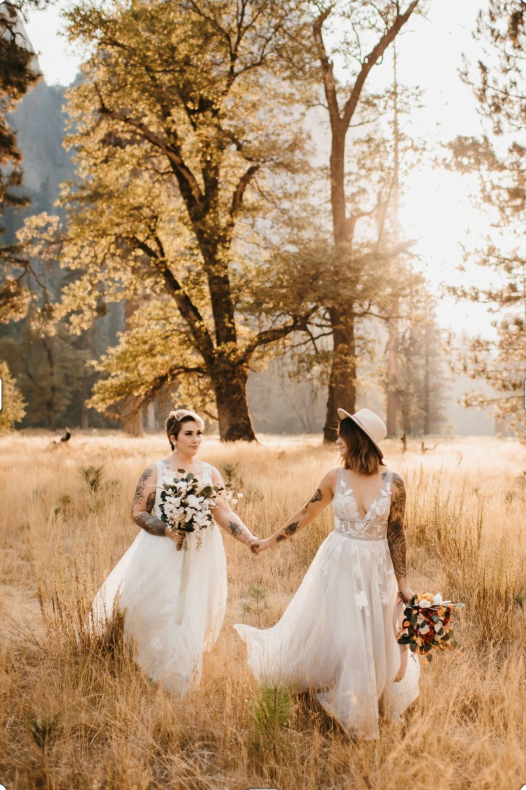 Newlyweds in white gowns walk hand in hand through autumn field with golden sunlight filtering through trees.