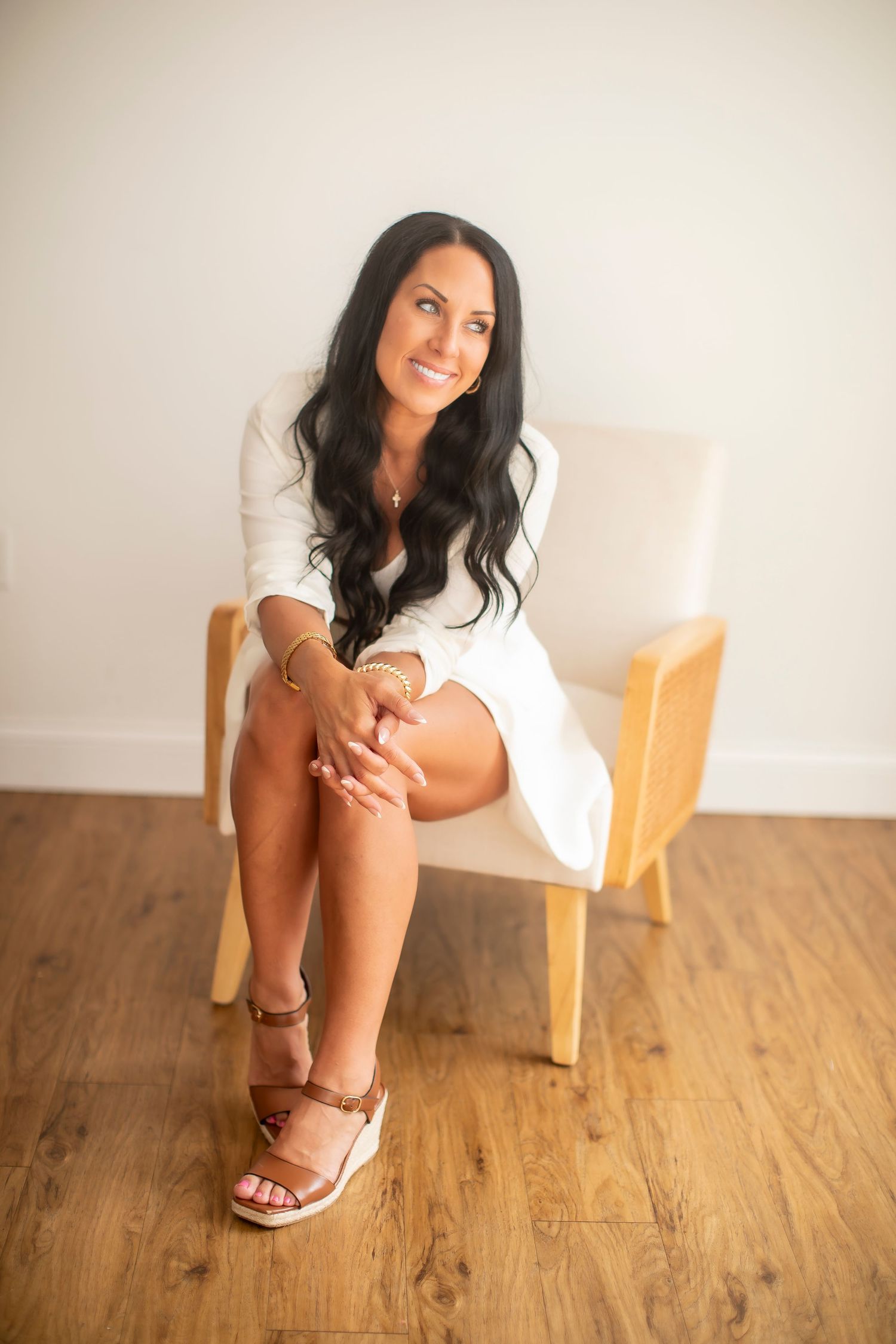Brunette woman in a white dress sitting in a chair on hardwood floors in a bright room.