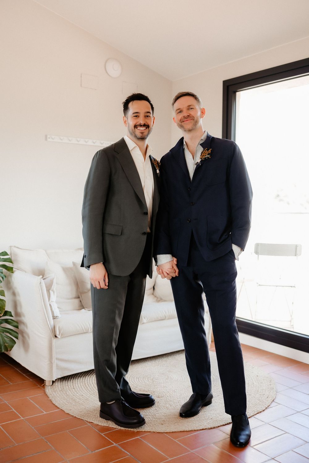Two people in formal attire stand together in a bright modern room with white furniture and terracotta flooring.