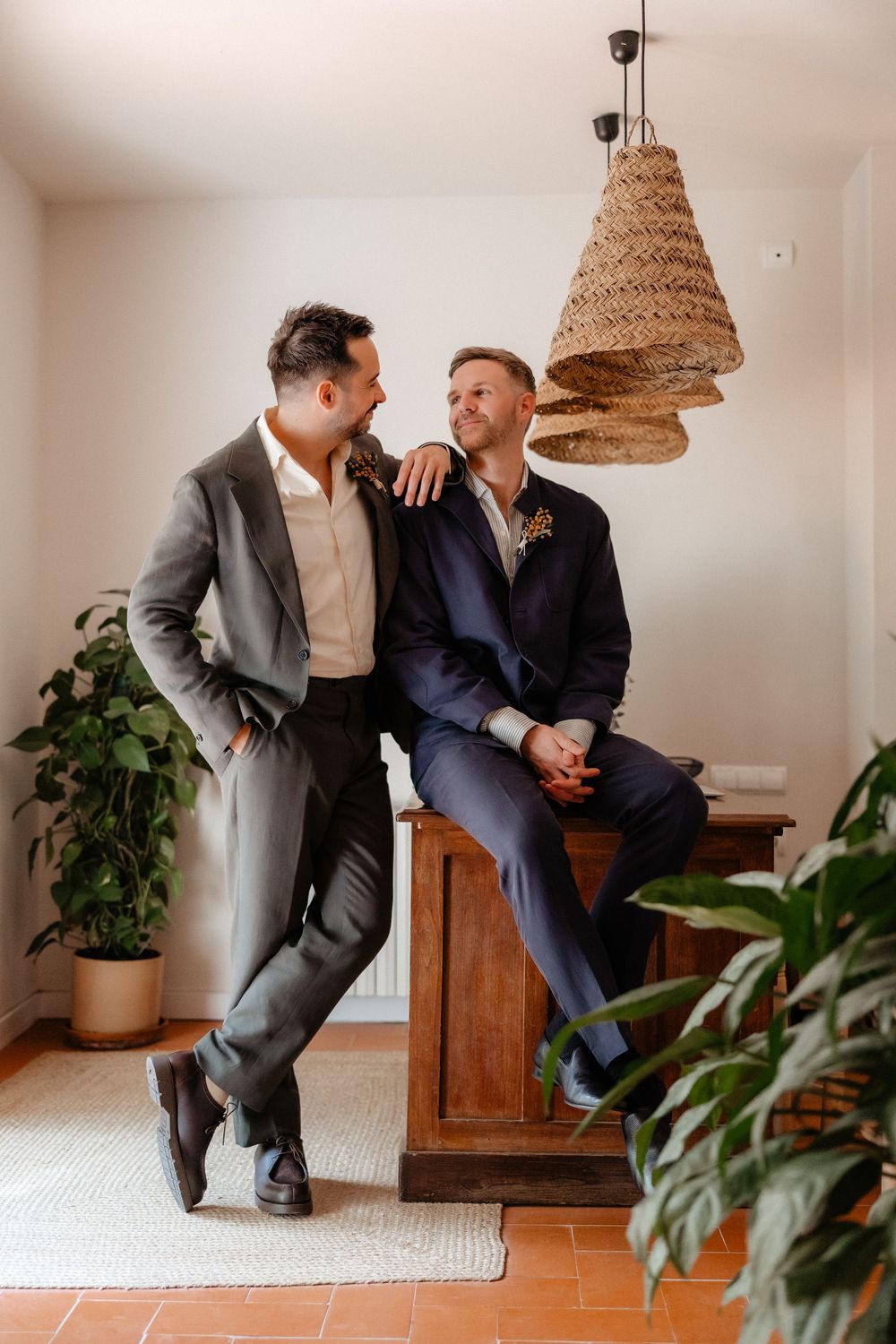 Two people in formal attire share an intimate conversation while seated on a wooden bench under woven pendant lights.