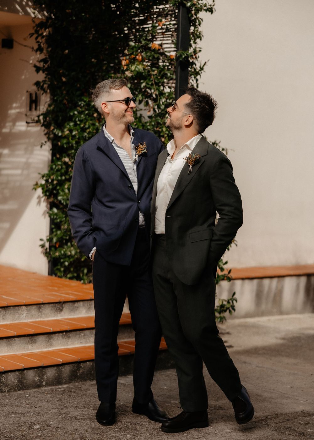 Two people in suits share a joyful moment together against a wall with climbing vines and wooden benches.