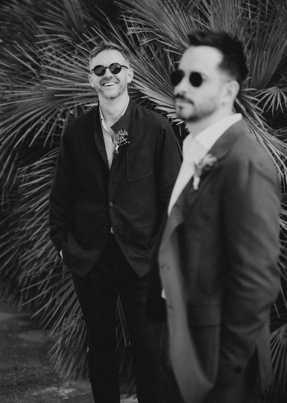 Two people in dark suits and sunglasses share a laugh while standing in front of palm fronds.
