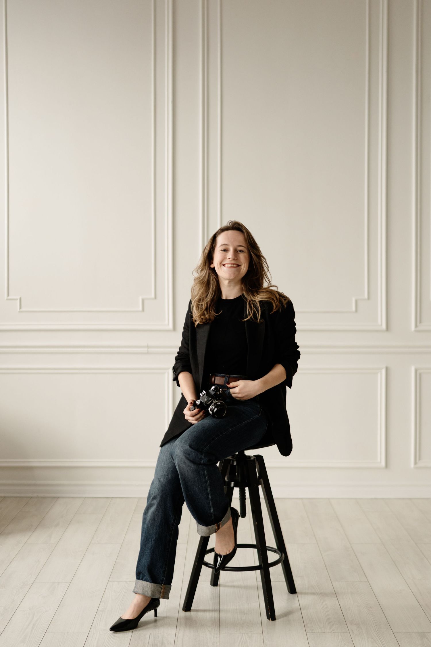 Four artistic portrait photos show a person in black attire posing on a wooden stool against a white wall.