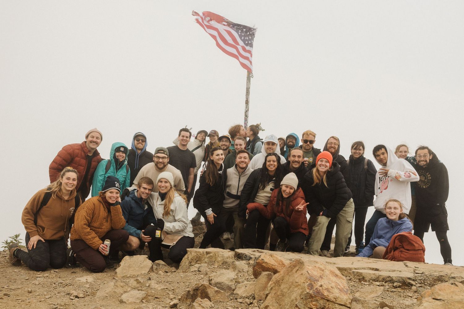 Large group of hikers posing together at a foggy mountain summit around an American flag pole.