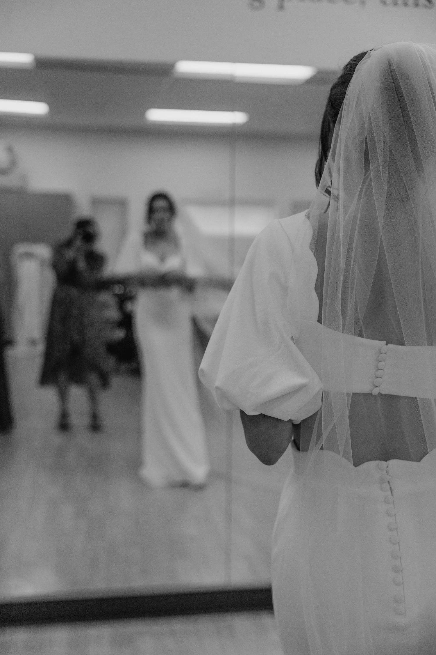Black and white photo of bride in white gown and veil preparing in a dressing room mirror.